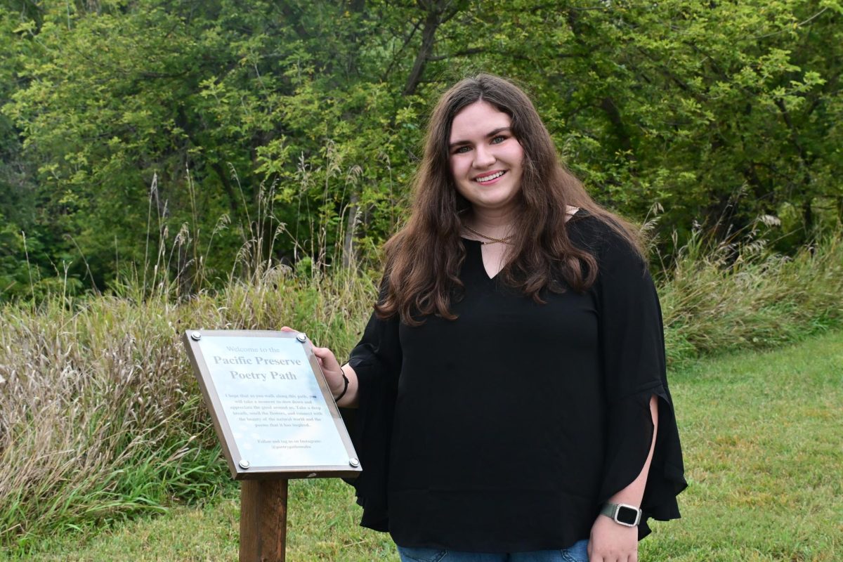 Senior Gabby Burns stands next to a sign reading "Welcome to the Pacific Poetry Path," celebrating the launch of the new trail featuring local poetry. The trail, part of her IB CAS project, displays poems from both professional and middle school poets. 