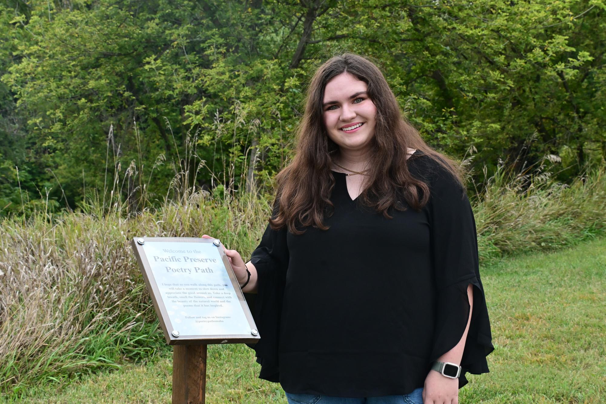Senior Gabby Burns stands next to a sign reading "Welcome to the Pacific Poetry Path," celebrating the launch of the new trail featuring local poetry. The trail, part of her IB CAS project, displays poems from both professional and middle school poets. 